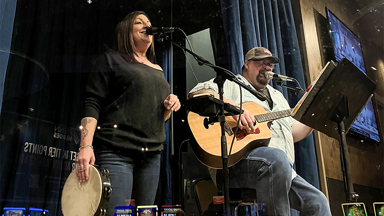 Man and woman music duo singing on stage with a microphone in center of the casino. Woman is playing the tambourine, and the man is playing an acoustic guitar. 