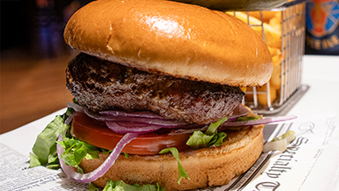 All American burger on brioche bun with newspaper liner. On a white plate with french fries and beer.